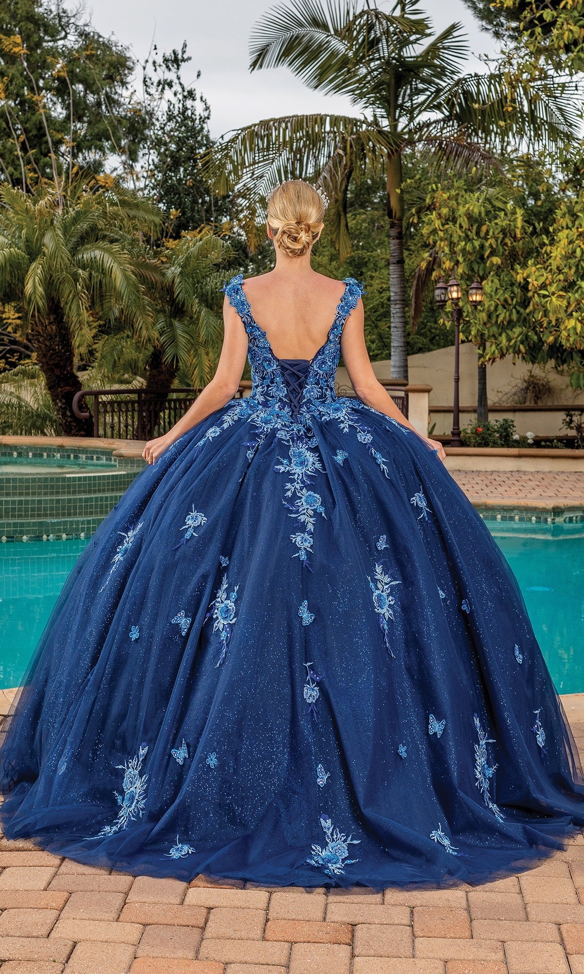 A woman with blonde hair in an updo wears the Quinceanera Dress 1886 By Dancing Queen, a navy ball gown with floral embroidery and a v-neckline, standing poolside with palm trees in the background.